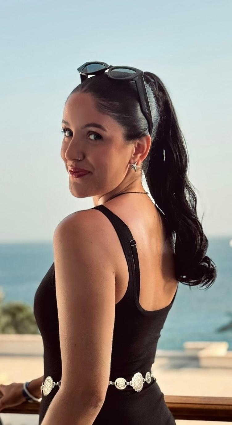 Femme aux cheveux foncés coiffés en une longue queue de cheval et portant un débardeur noir et des lunettes de soleil sur la tête, souriant avec la mer derrière elle.