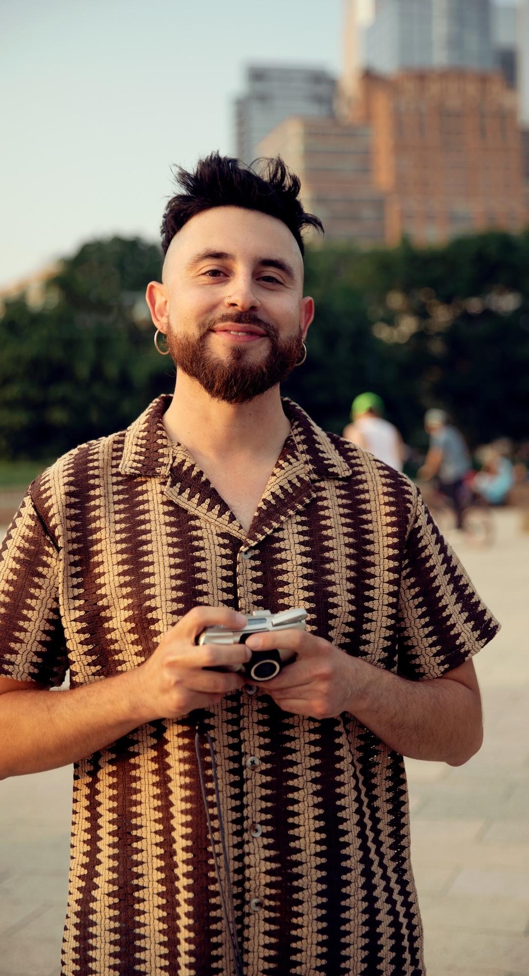 Homme barbu portant une chemise rayée, tenant un appareil photo, et se tenant debout, à l’extérieur, avec des arbres et des bâtiments en arrière-plan.